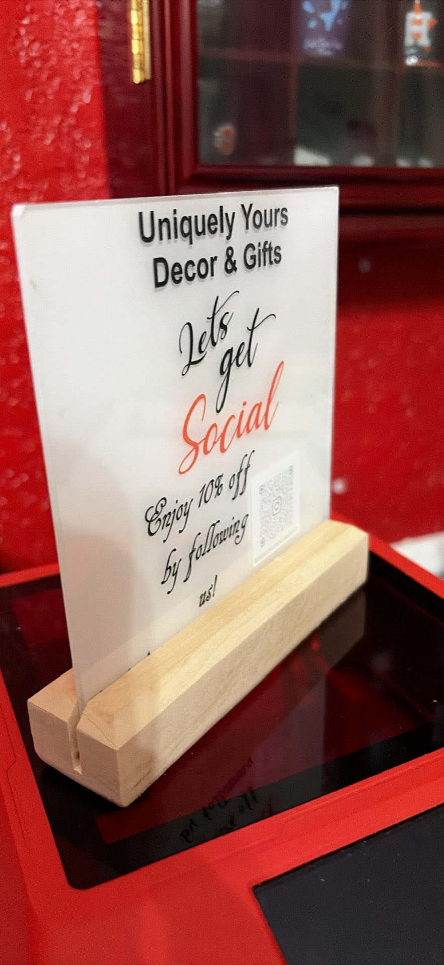 A white printed sign with black and red text reading 'Uniquely Yours Decor & Gifts' and 'Let's get Social', displayed on a small light wood stand on a red surface.