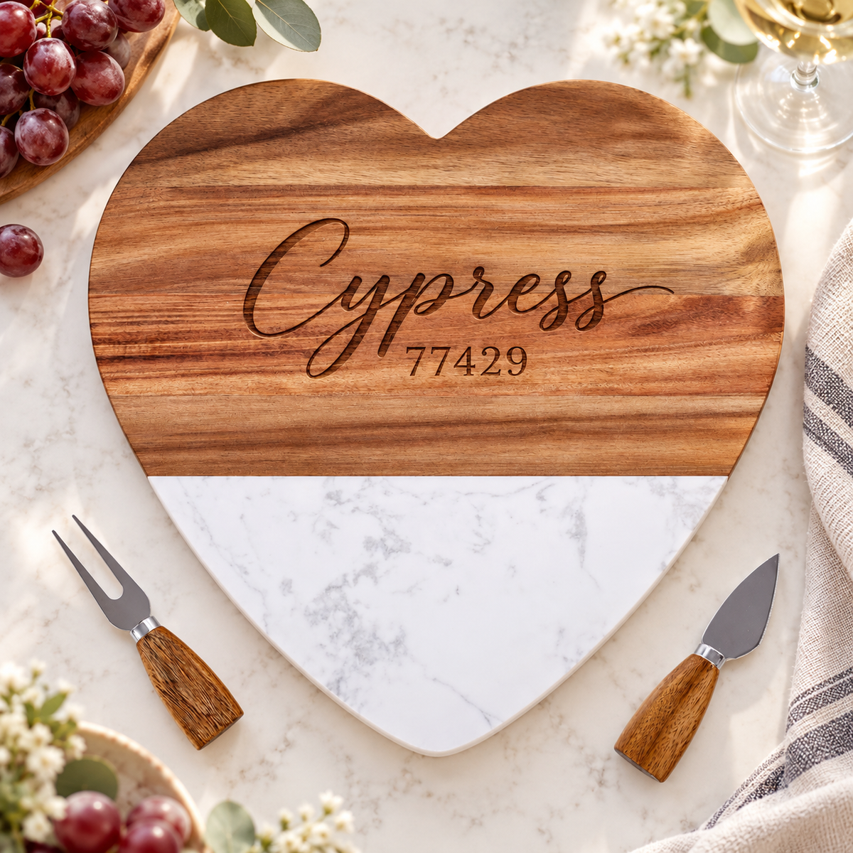 Marble and wood heart\-shaped cheese board with two wood\-handled serving utensils, available with personalized names or initials\.