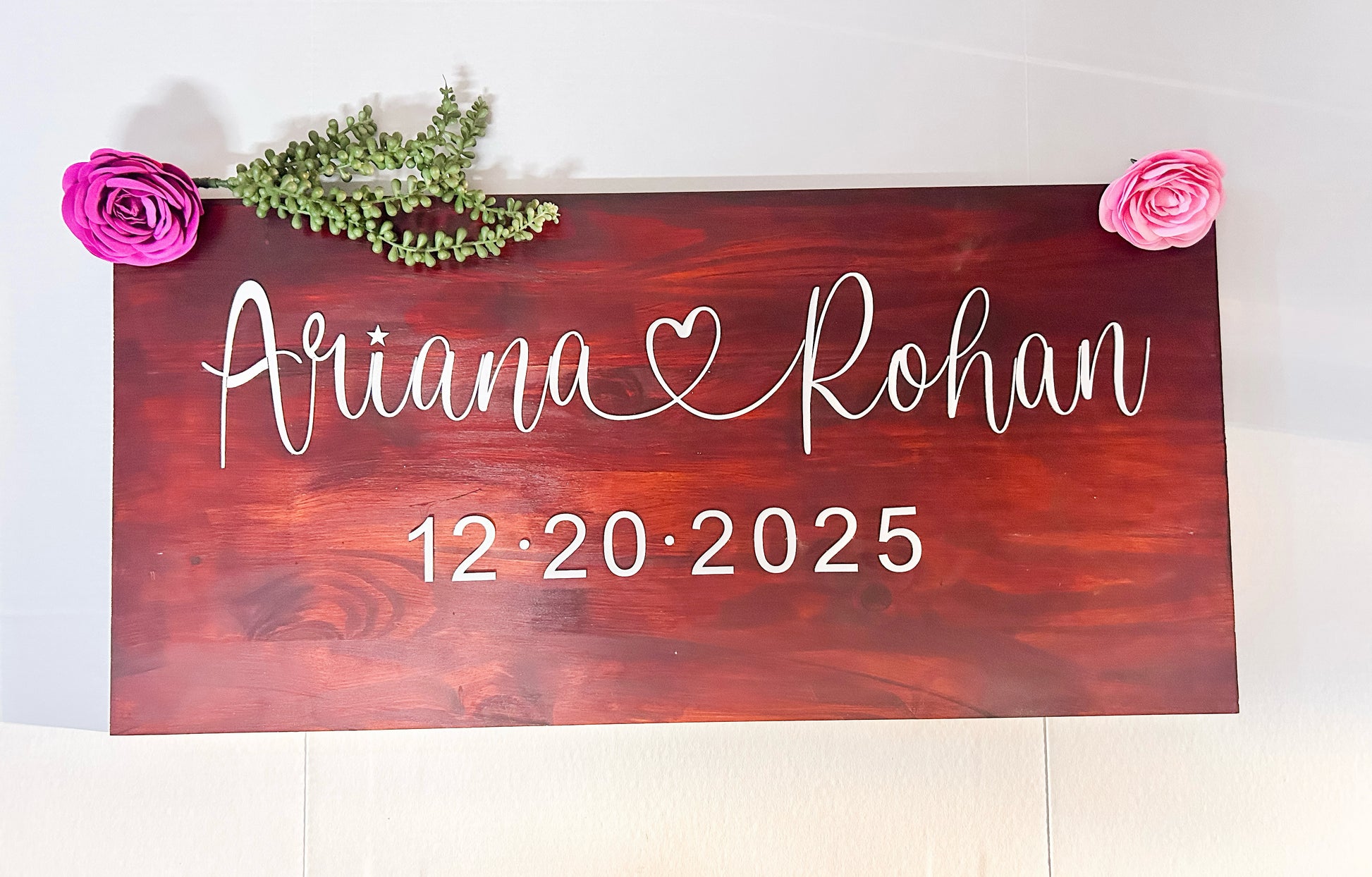 Handcrafted wooden wedding sign adorned with pink roses, ivory flowers, and greenery garland. Personalized wedding decor featuring couple names and wedding date for ceremony and reception display.